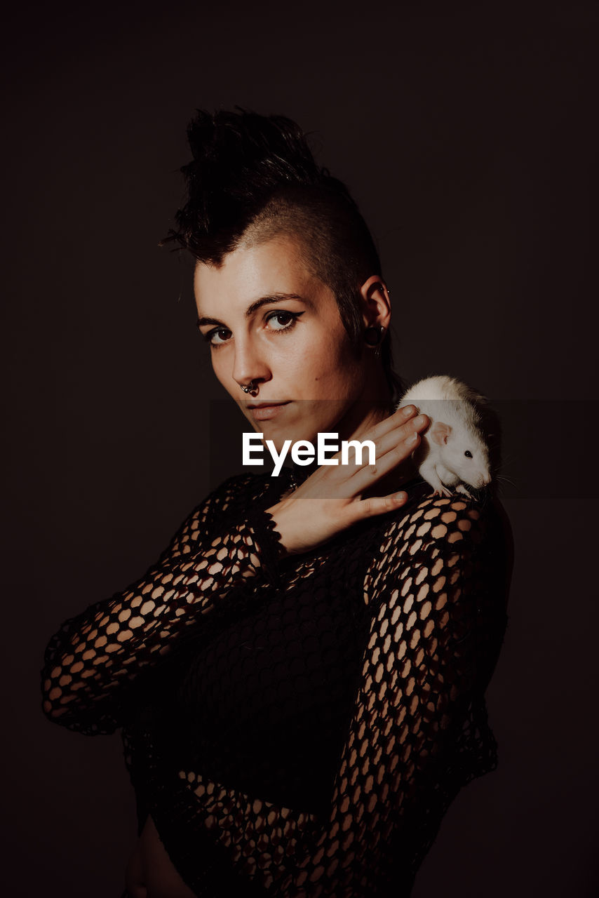 Confident woman with mohawk carrying white rat on shoulder and looking at camera against black background