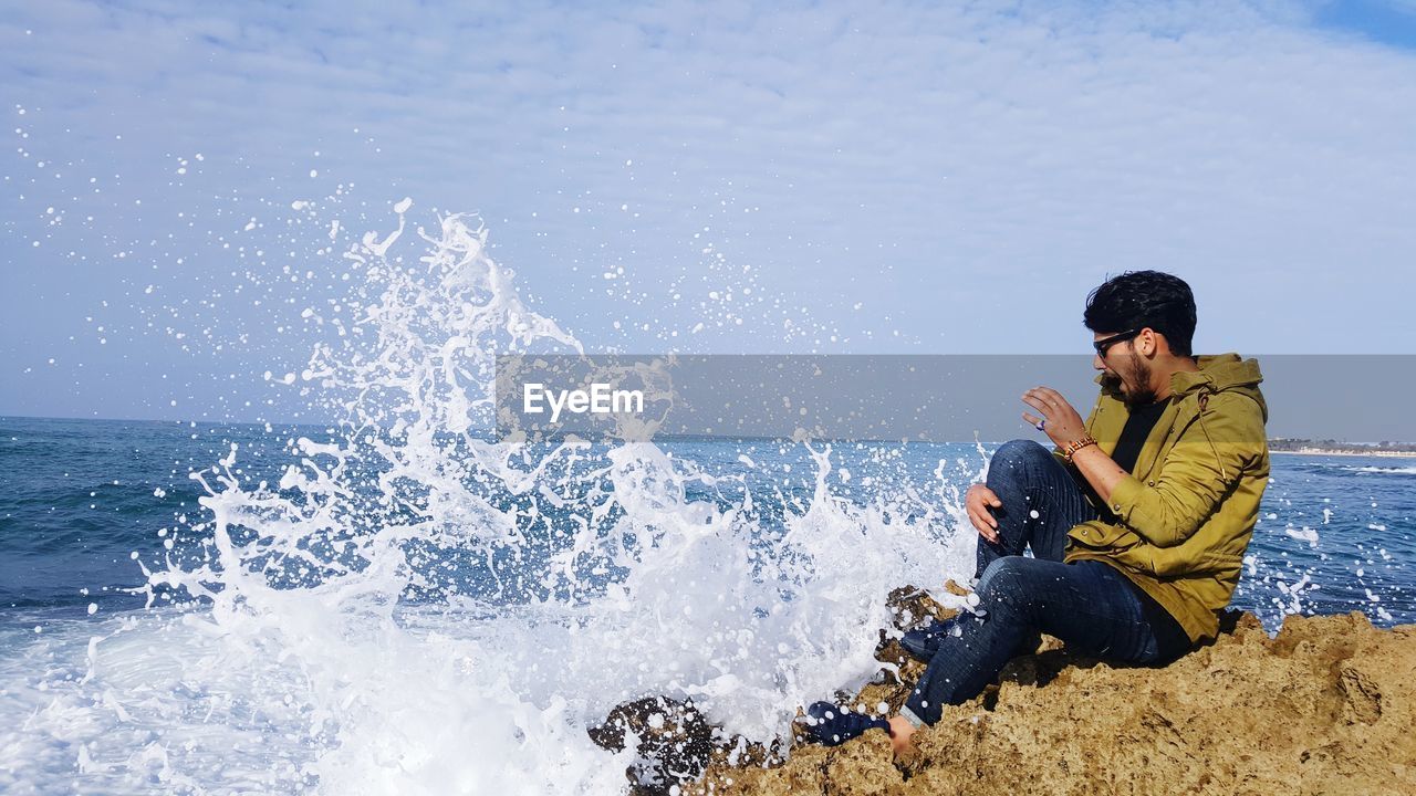 Side view of young man sitting on rock by sea against cloudy sky