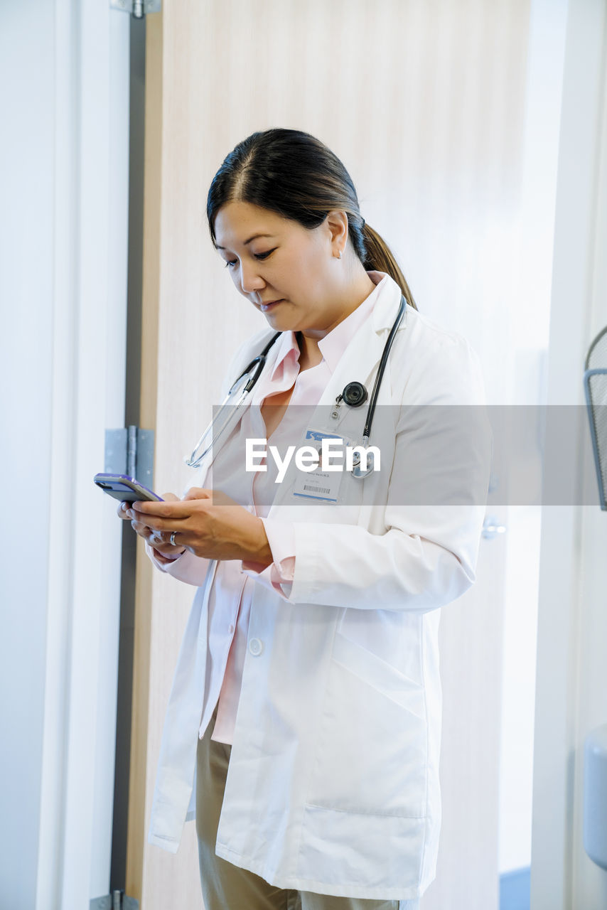 Female doctor using smart phone while standing against door in hospital