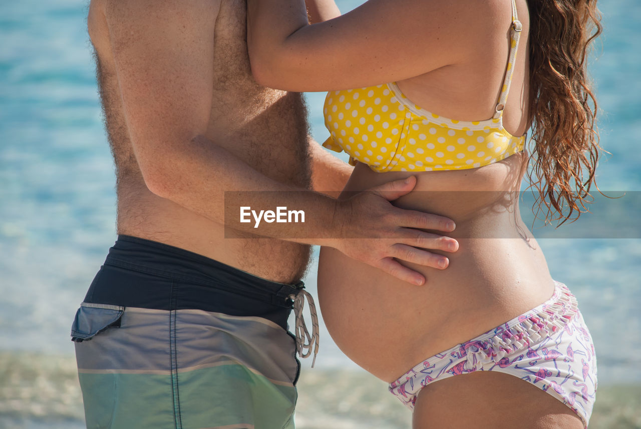 Midsection of pregnant woman in bikini and husband standing by water