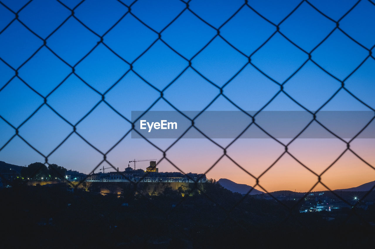 Silhouette cityscape behind chain link fence against sky during sunset