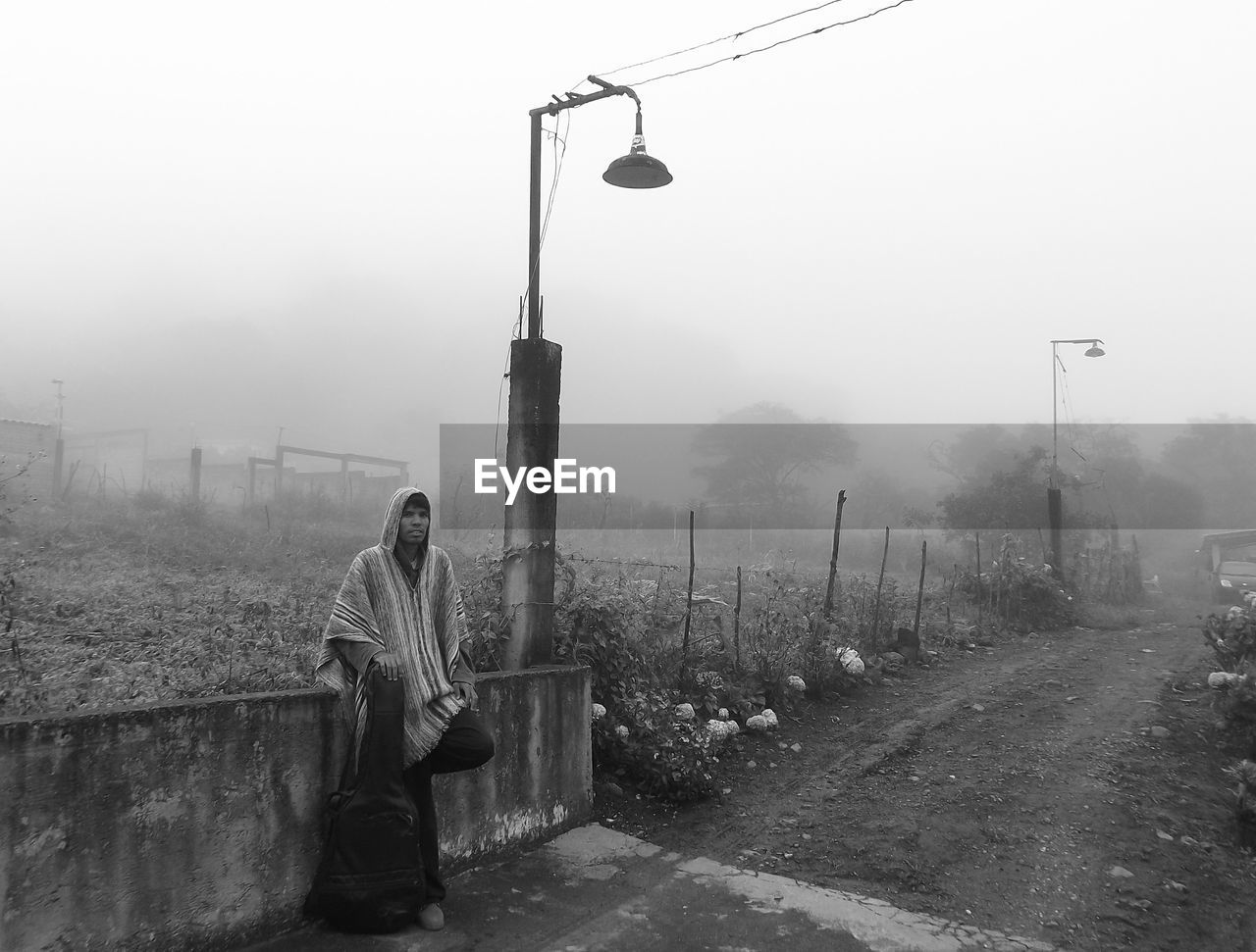 Full length of young man with guitar leaning on retaining wall during foggy weather