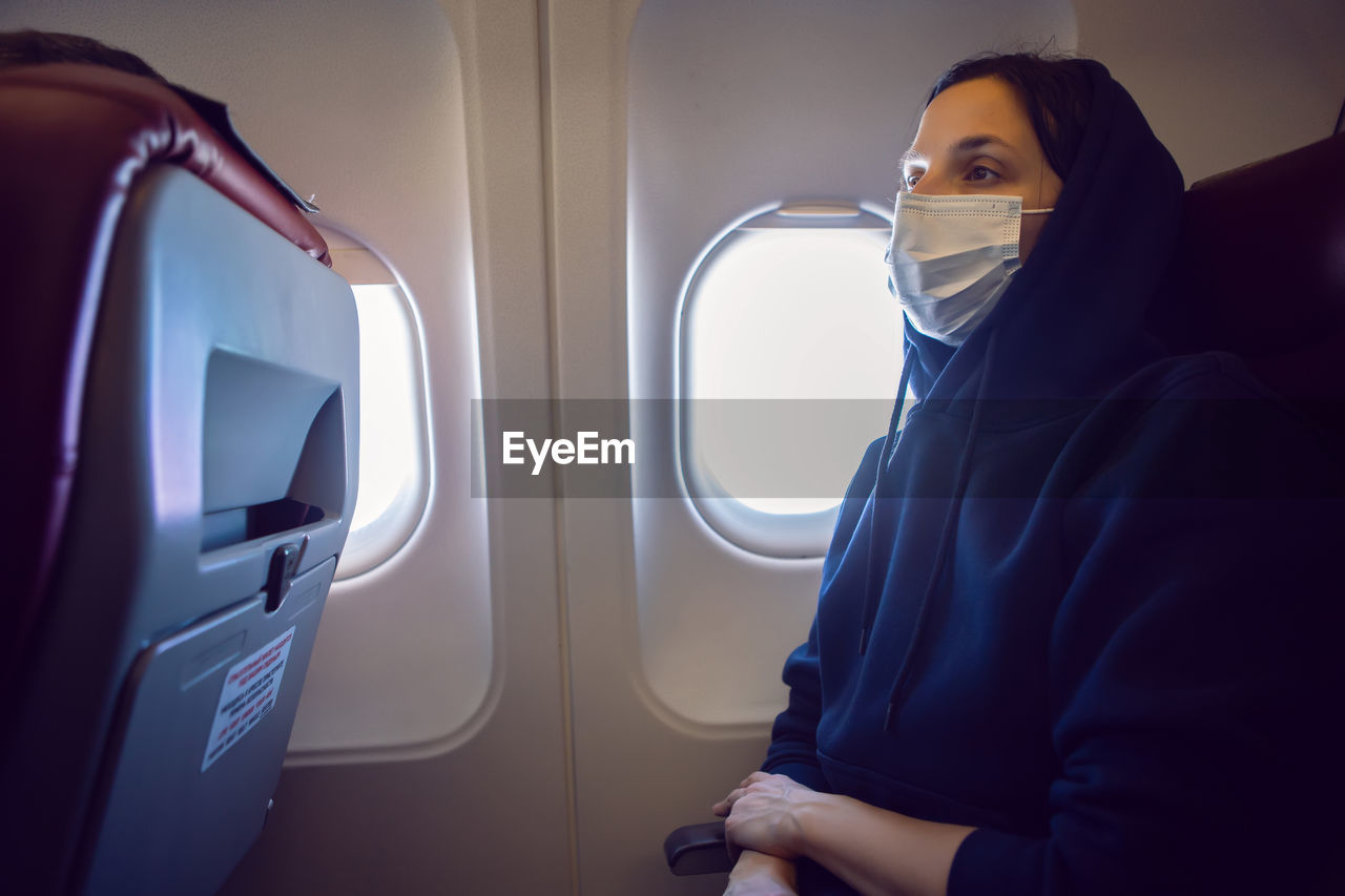 Woman in a black jacket with mask is a passenger of the plane sitting on chair next to the window