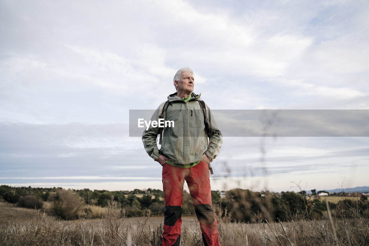 Senior male hiker with hands in pockets standing on agricultural field against cloudy sky at countryside