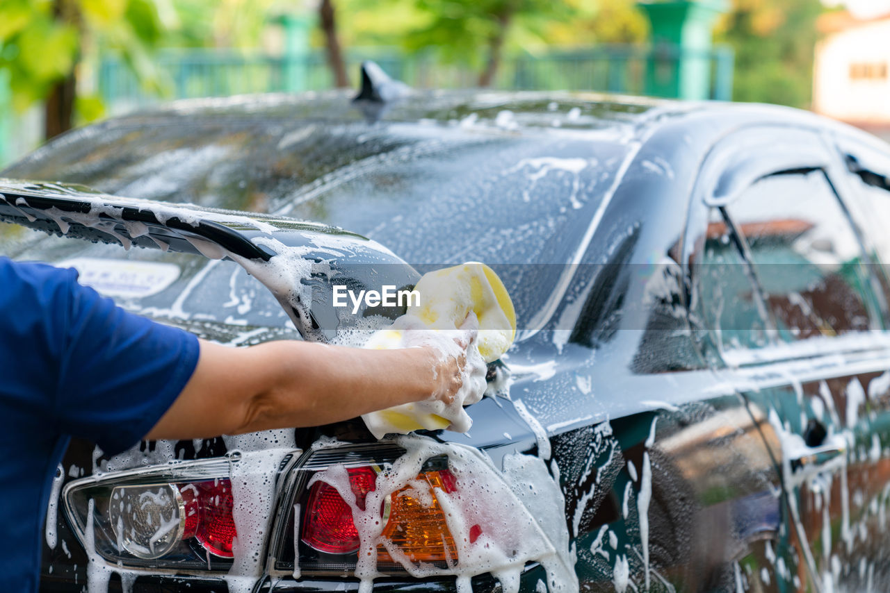 Woman hand hold yellow sponge washing spoiler black car at home. car detailing.