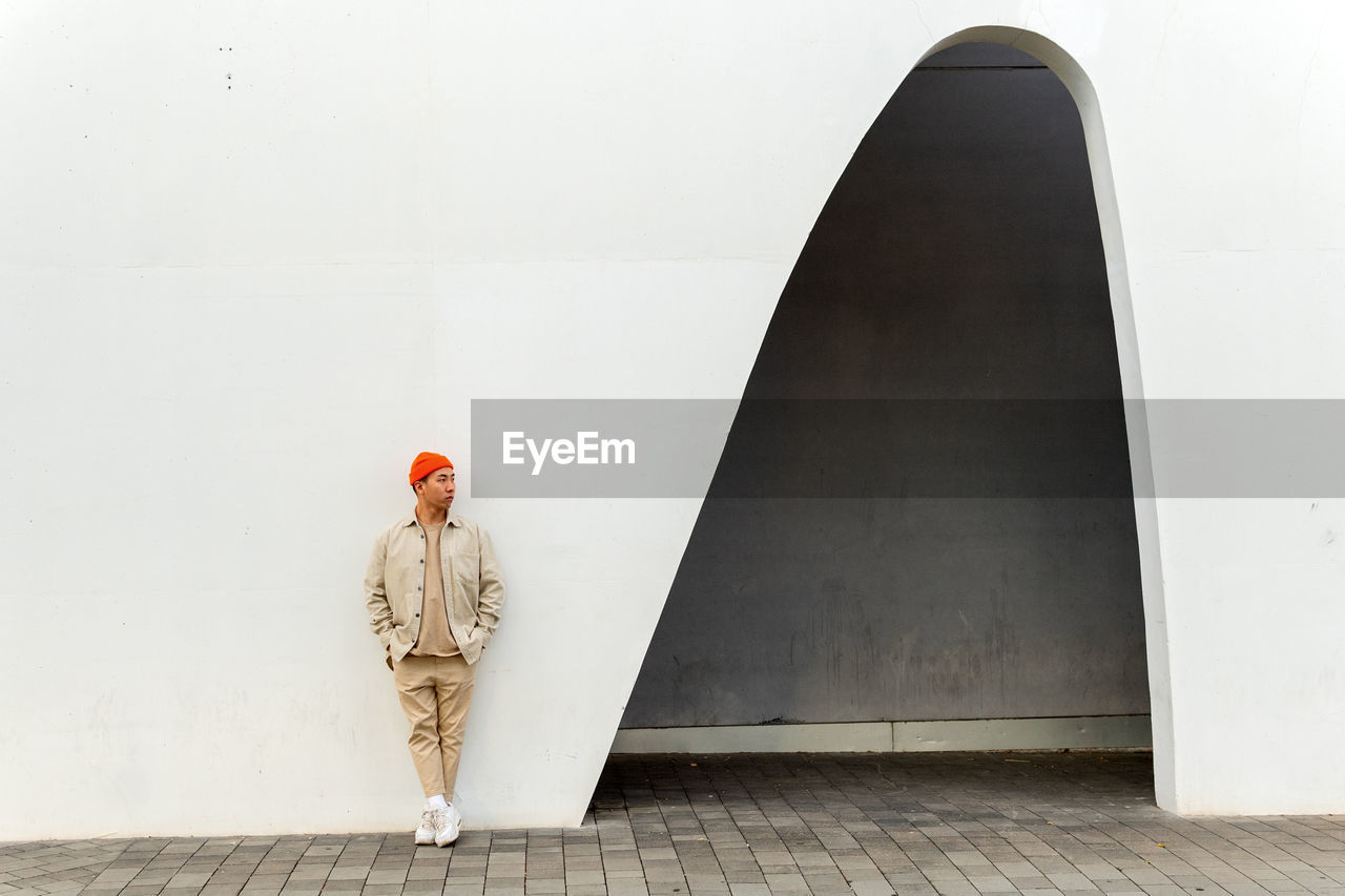 Full length thoughtful asian male wearing casual clothes and orange hat leaning with hands in pockets on white building wall with creative arch