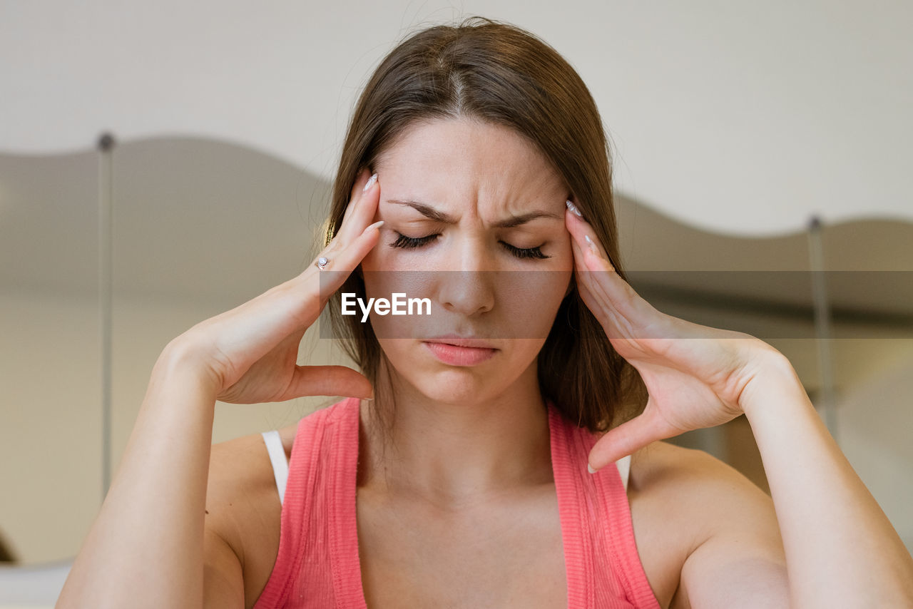 Young woman sitting on bed holding her head with her hands, suffering from