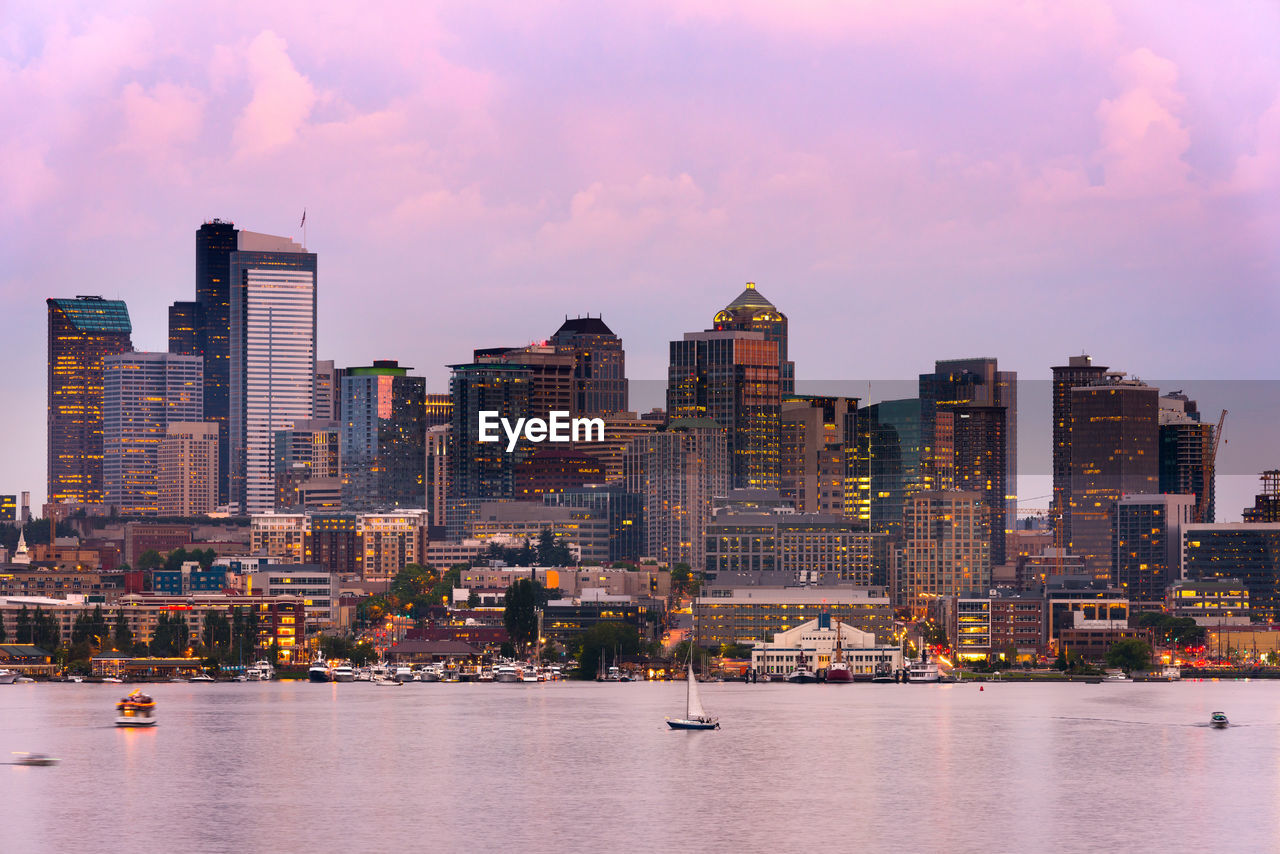 Modern buildings by river against sky during sunset