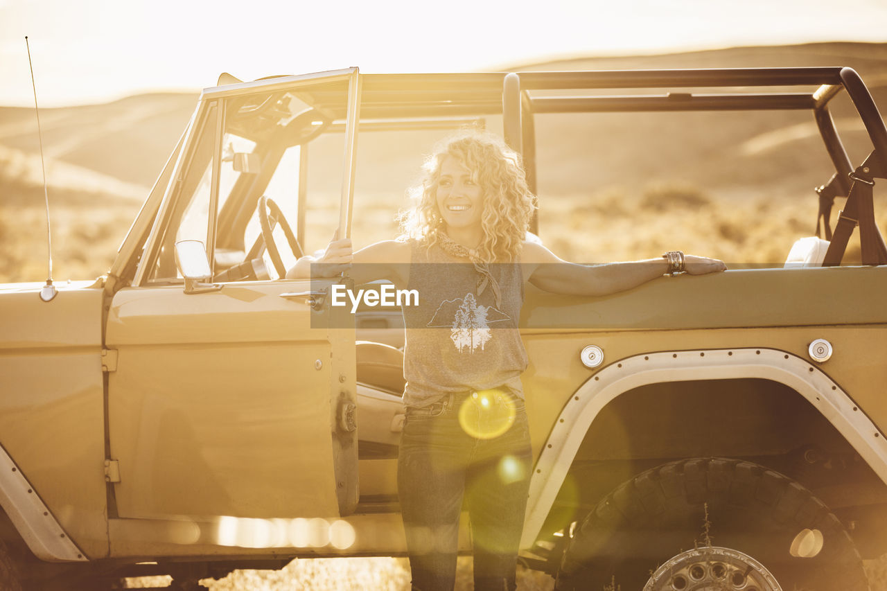 Cheerful woman looking away while standing against off-road vehicle during sunny day