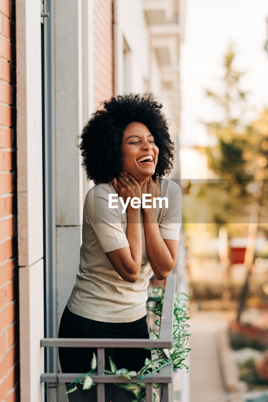 Happy african american female with eyes closed in casual outfit while standing on balcony in daytime