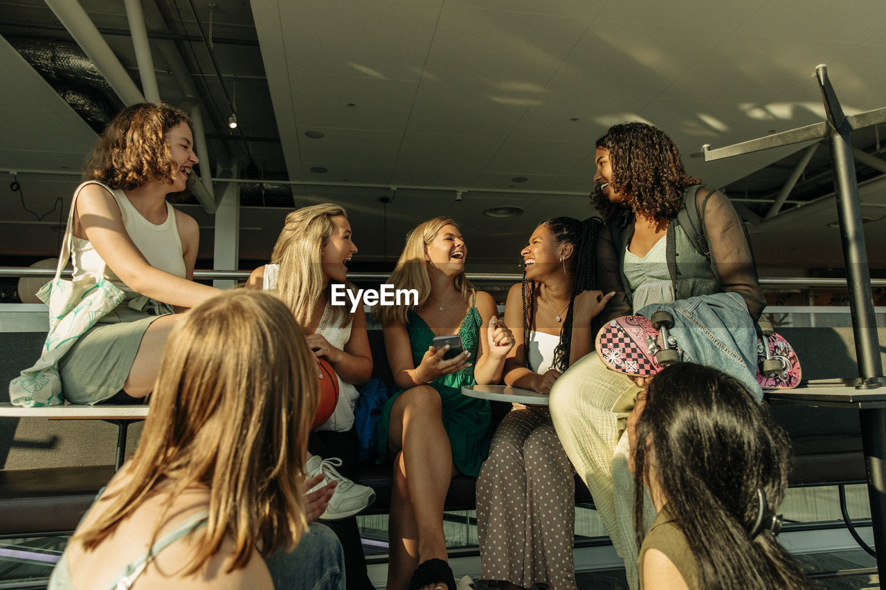 Cheerful teenage girls talking with each other while sitting outside mall during sunny day