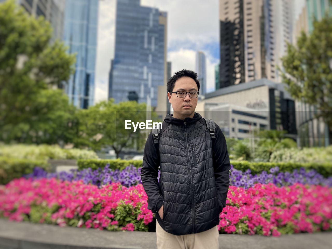Young asian man standing against flower beds, trees and skyscrapers in the city.