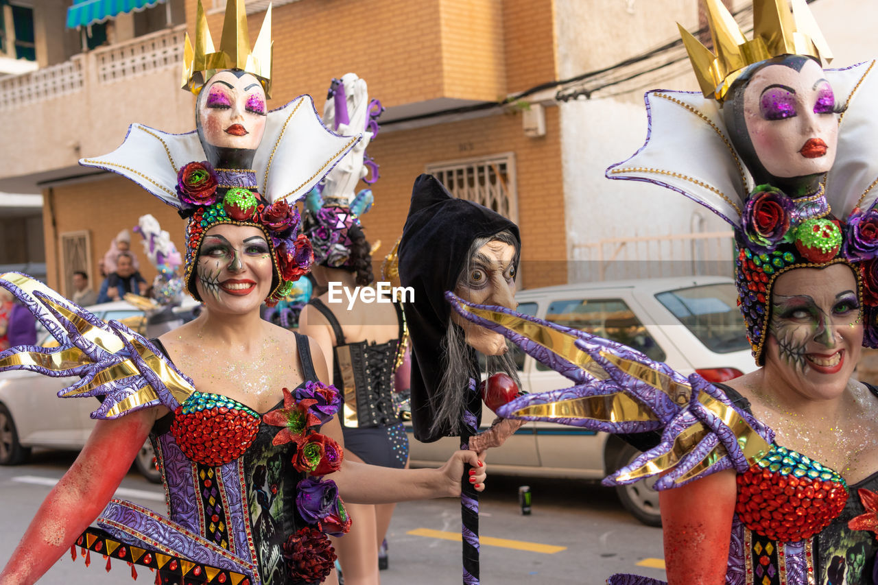 celebration, carnival, group of people, arts culture and entertainment, women, event, adult, traditional clothing, tradition, dancing, clothing, headwear, performance, fun, costume, smiling, architecture, happiness, young adult, enjoyment, headdress, togetherness, multi colored, portrait, city, emotion, disguise, festival, performing arts, friendship, lifestyles, mask - disguise, leisure activity, outdoors, fashion, person, female, men, mask, waist up
