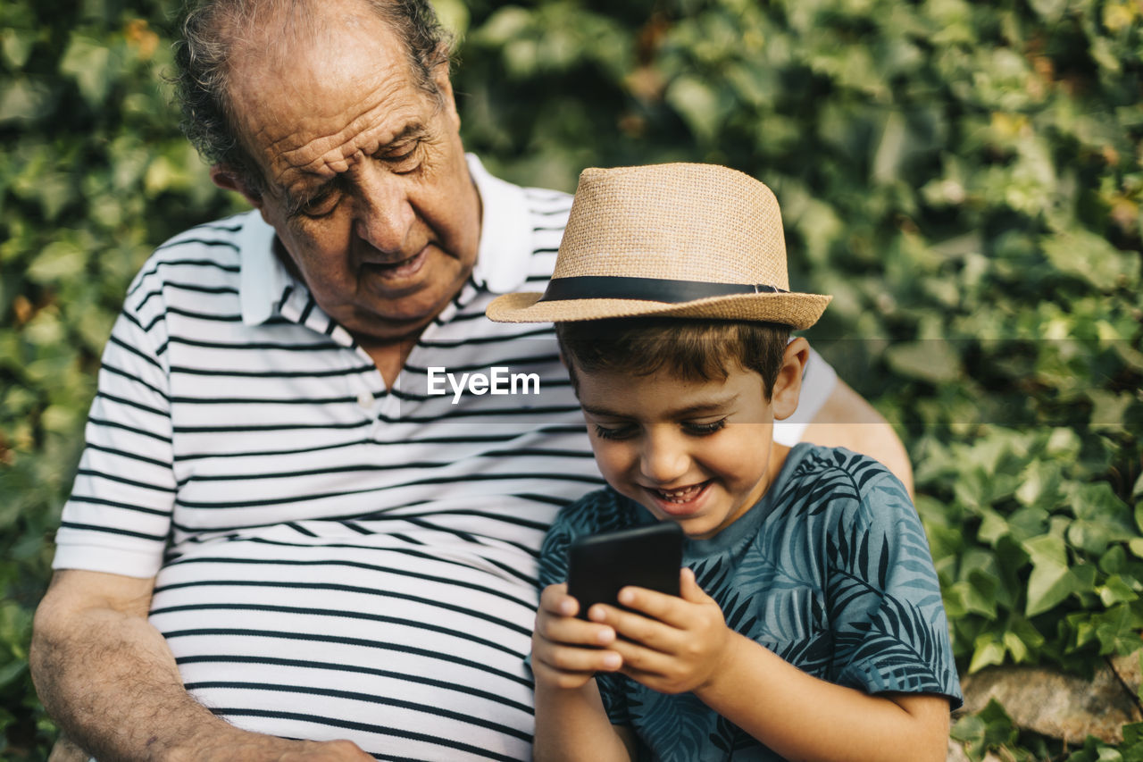 Cute boy wearing hat using smart phone while sitting with grandfather outdoors