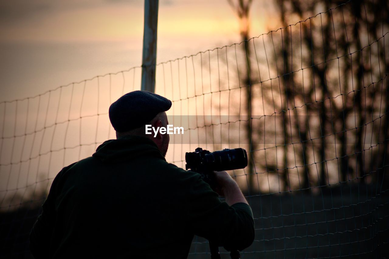 Rear view of man photographing while standing by fence against sky during sunset