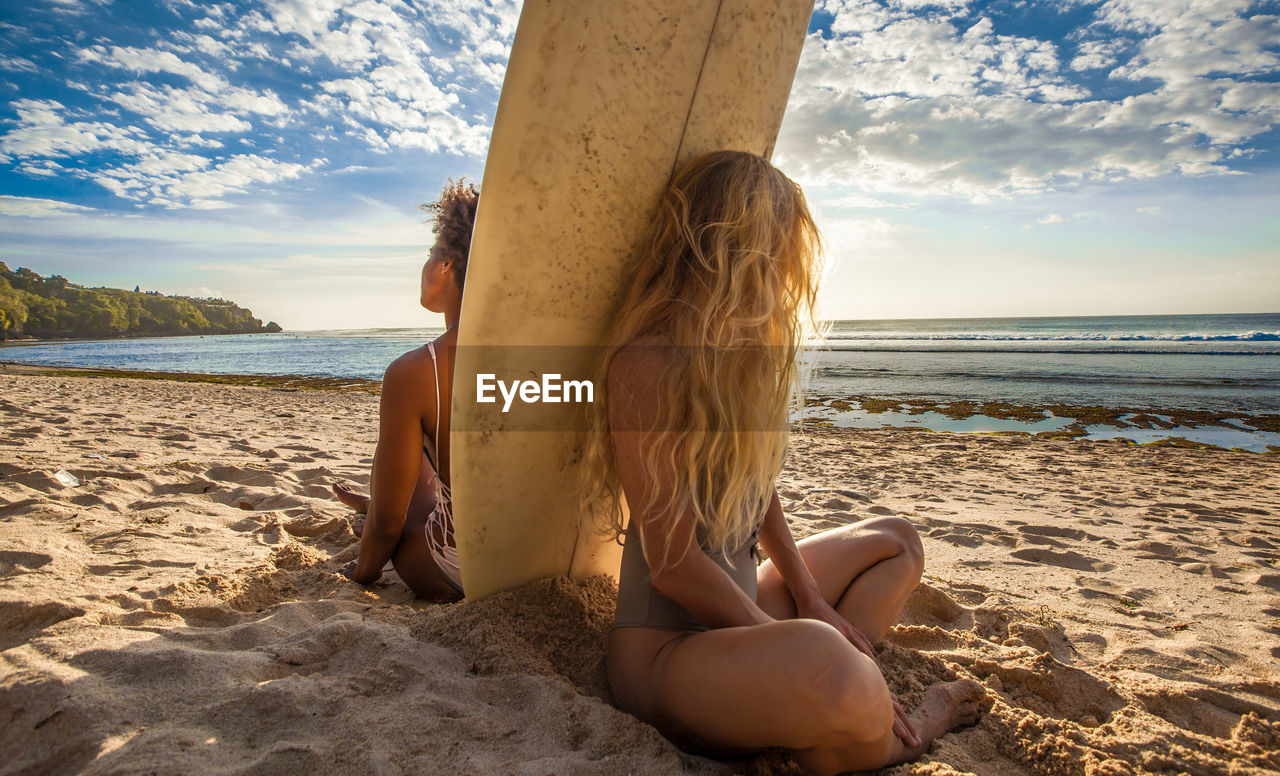 Friends with surfboard sitting on sand at beach against sky