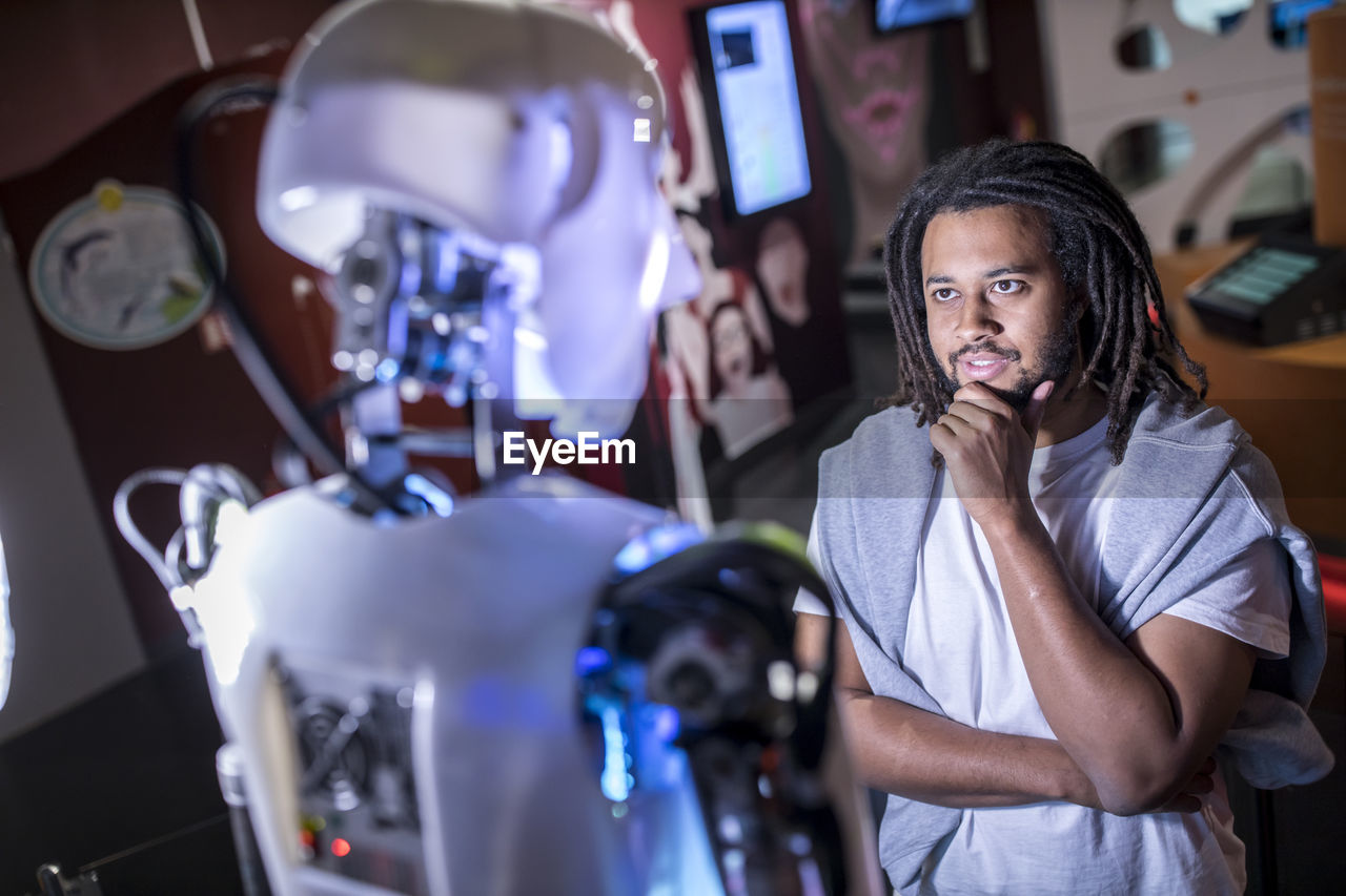 Thoughtful technician with hand on chin looking at human robot in workshop