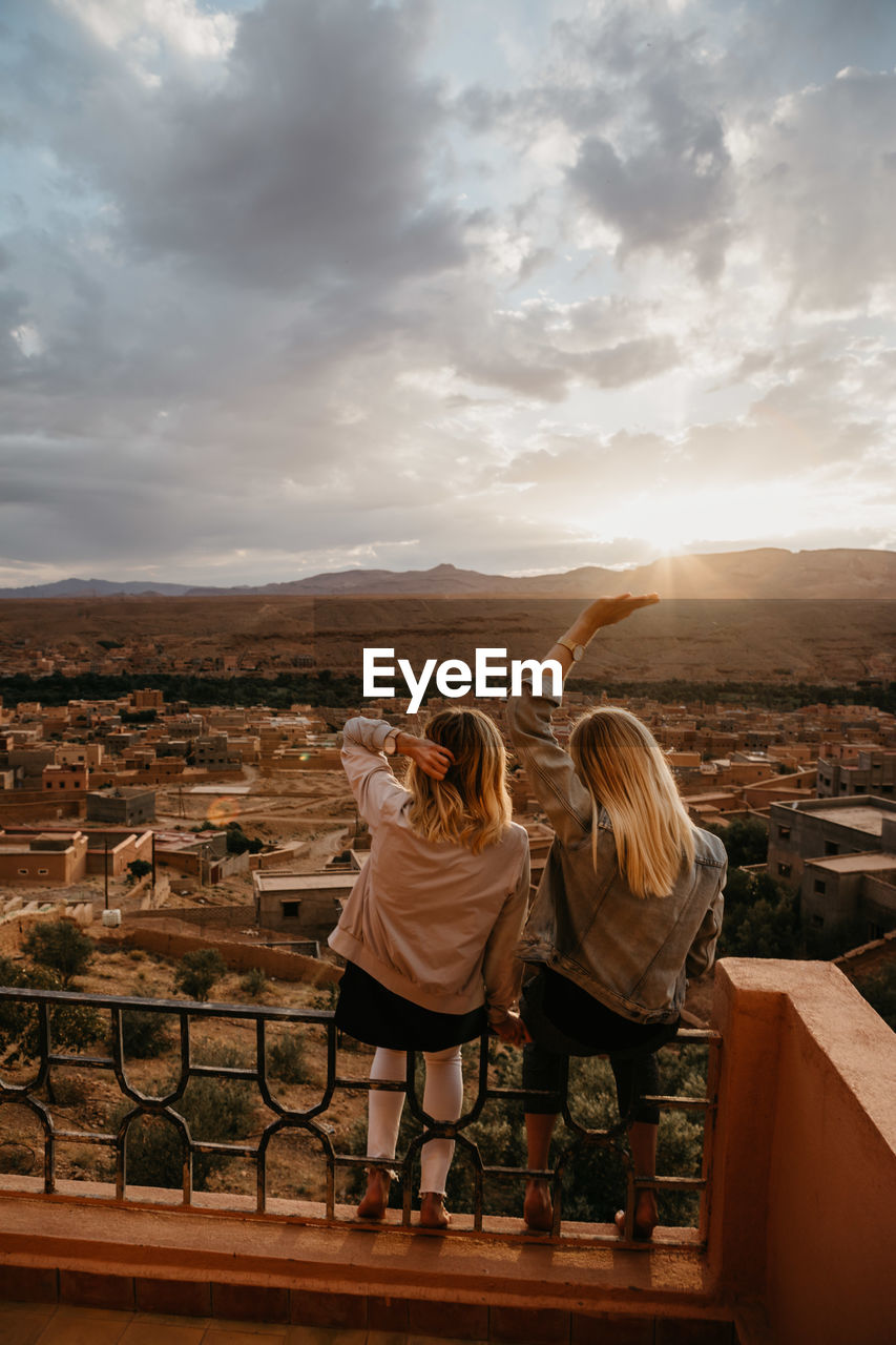 REAR VIEW OF WOMEN STANDING ON LANDSCAPE AGAINST SKY DURING SUNSET