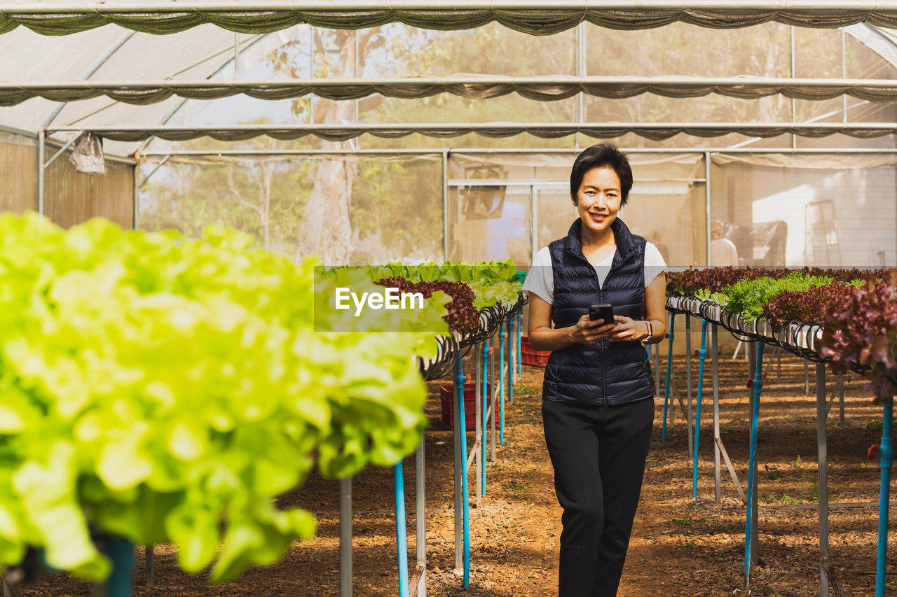 Portrait of happy woman farmer standing in organic vegetable farm and using smart phone.