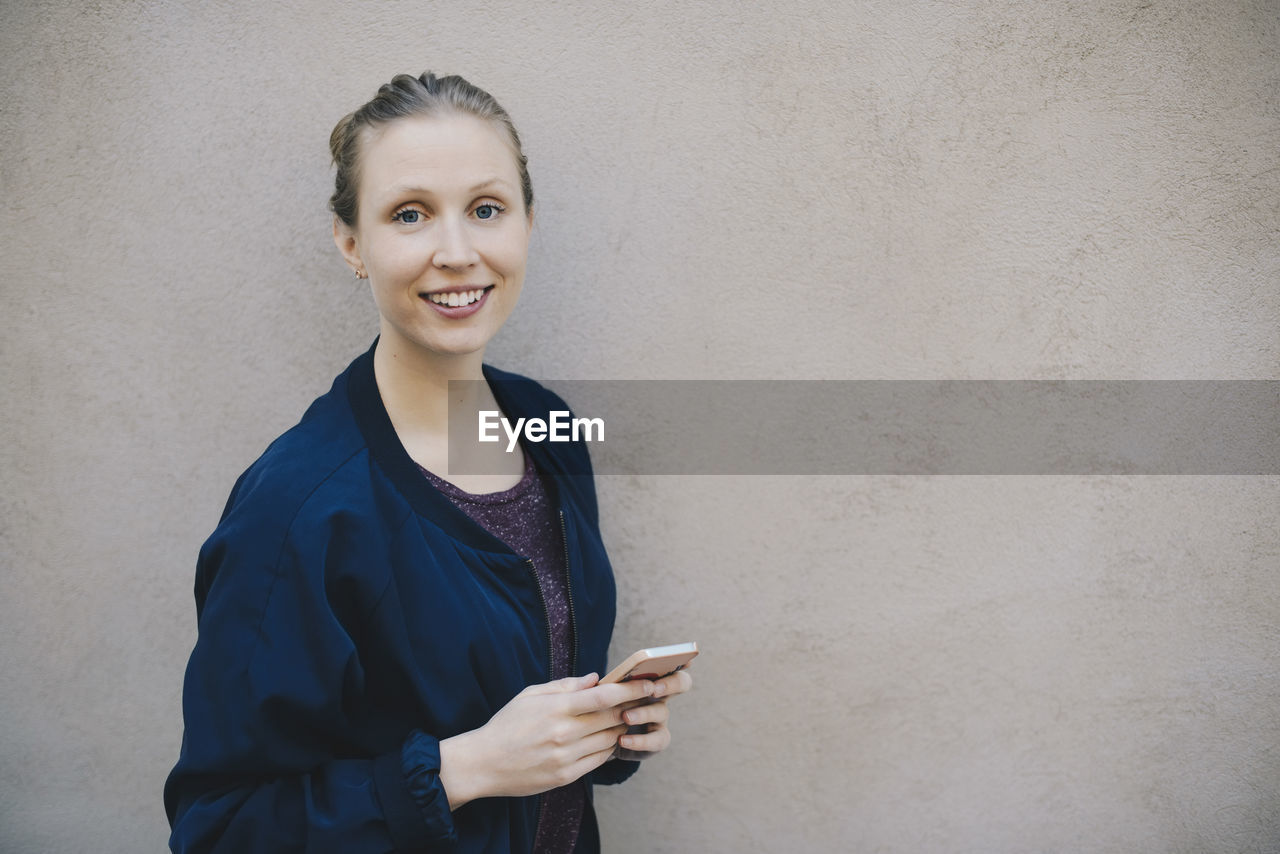 Portrait of happy female computer programmer holding smart phone while standing against beige wall in office