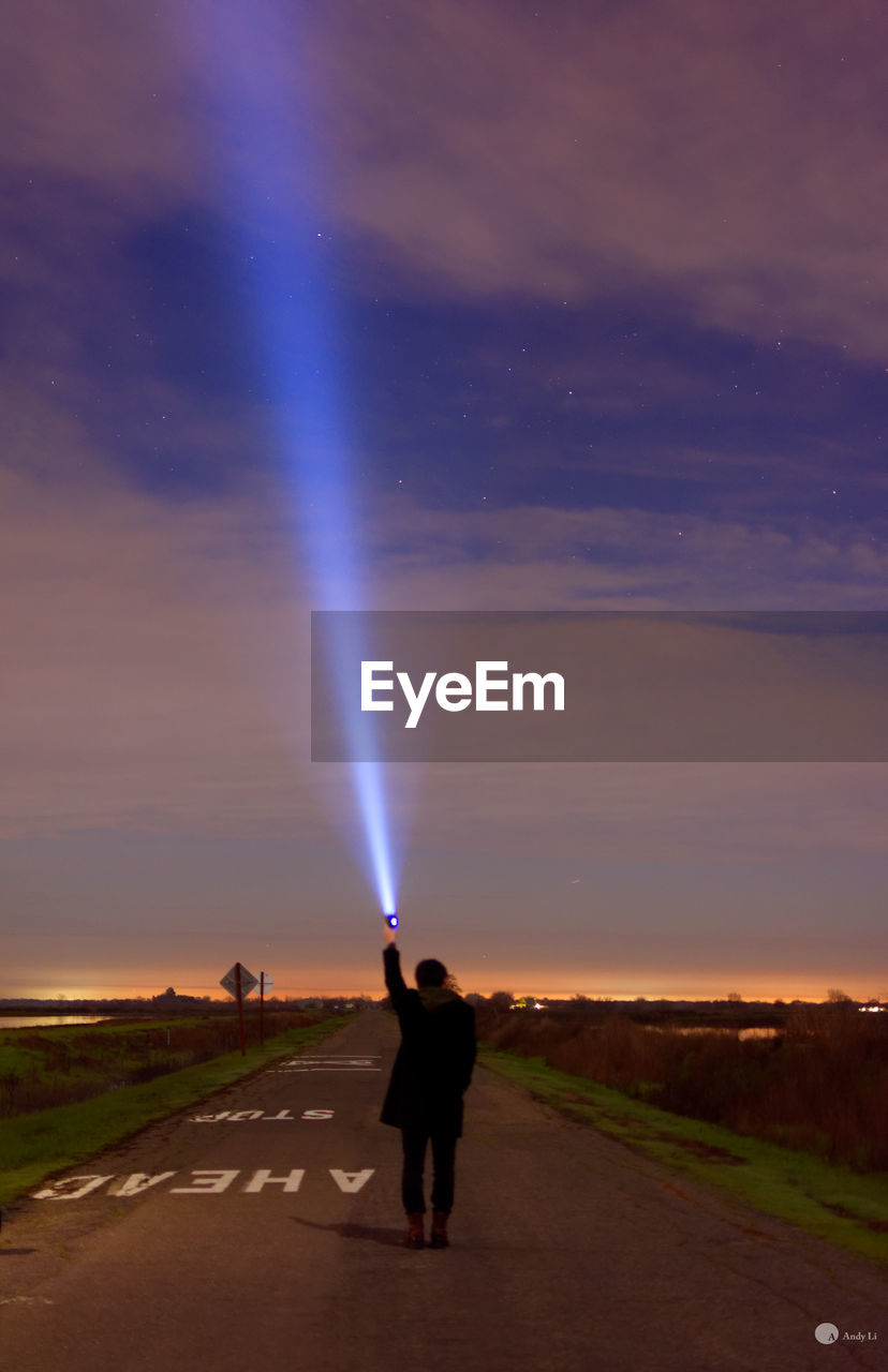 Rear view of person holding illuminated flashlight on road against cloudy sky at night