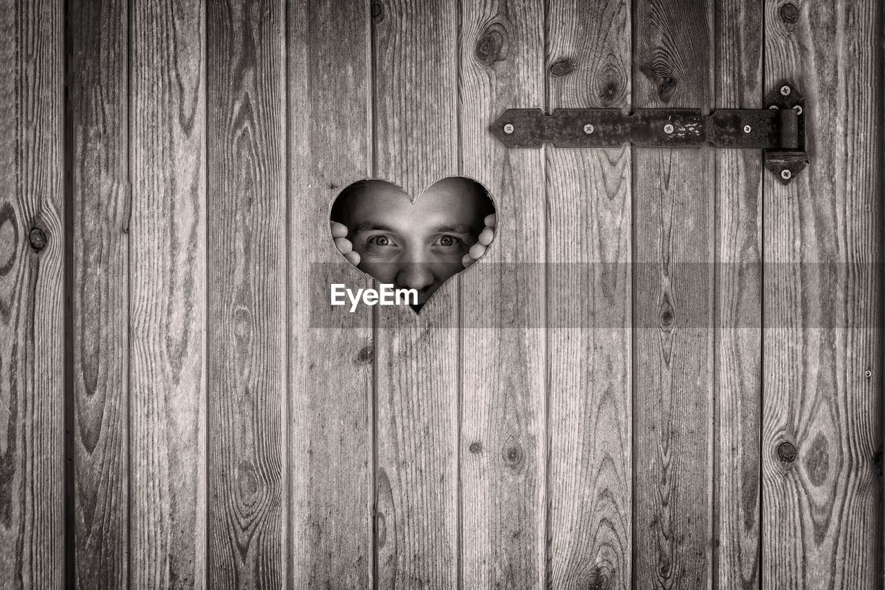 Close-up portrait of man peeking from heart shape on wooden door