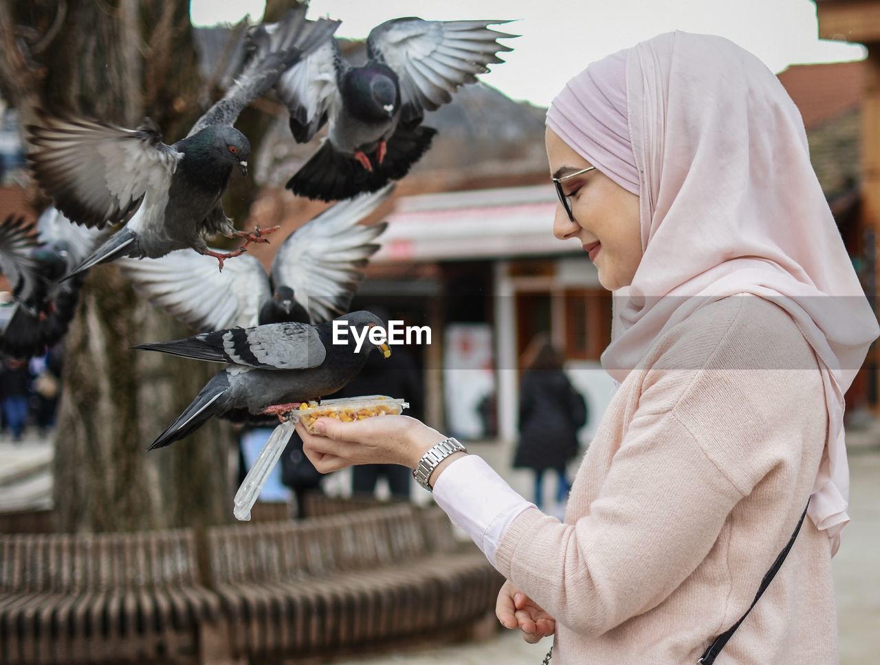 Side view of young woman feeding birds while standing on city street