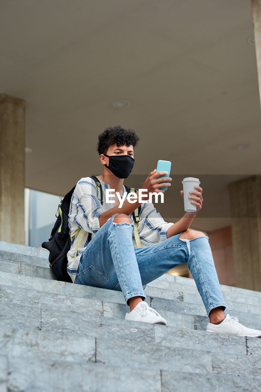 Young afro with mask uses his phone while he is sitting on the stairs