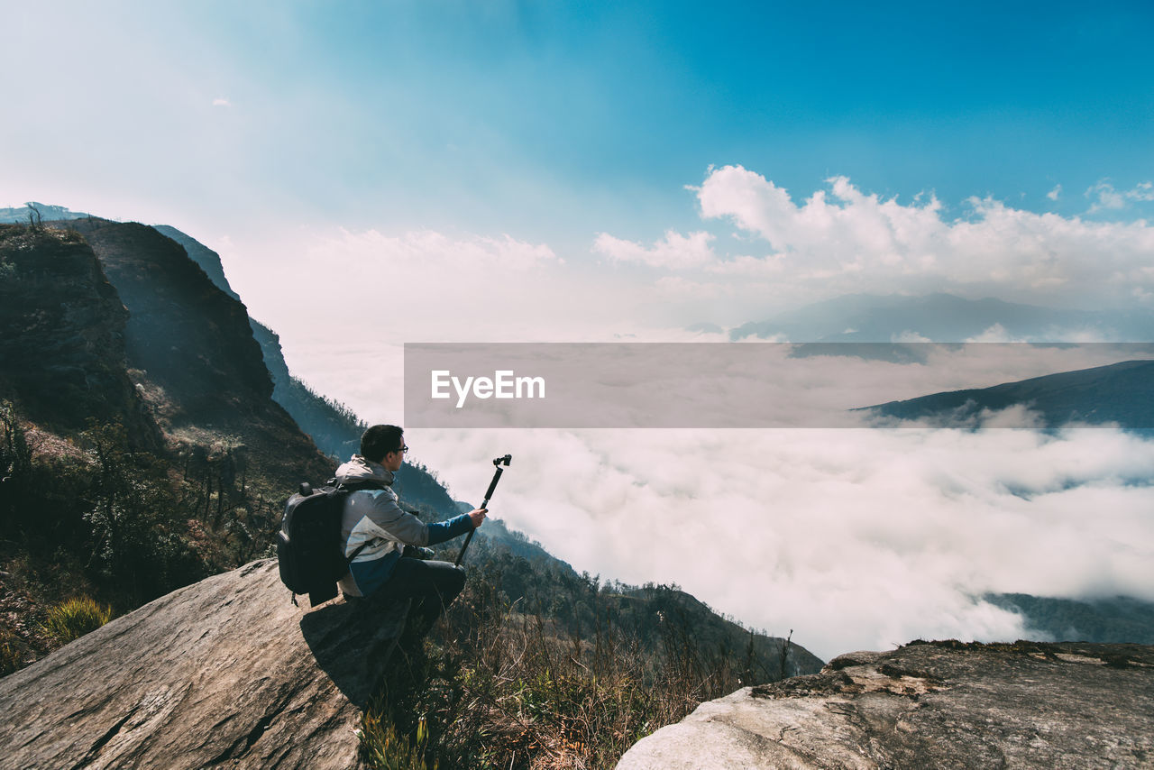 Man sitting on cliff against cloudy sky