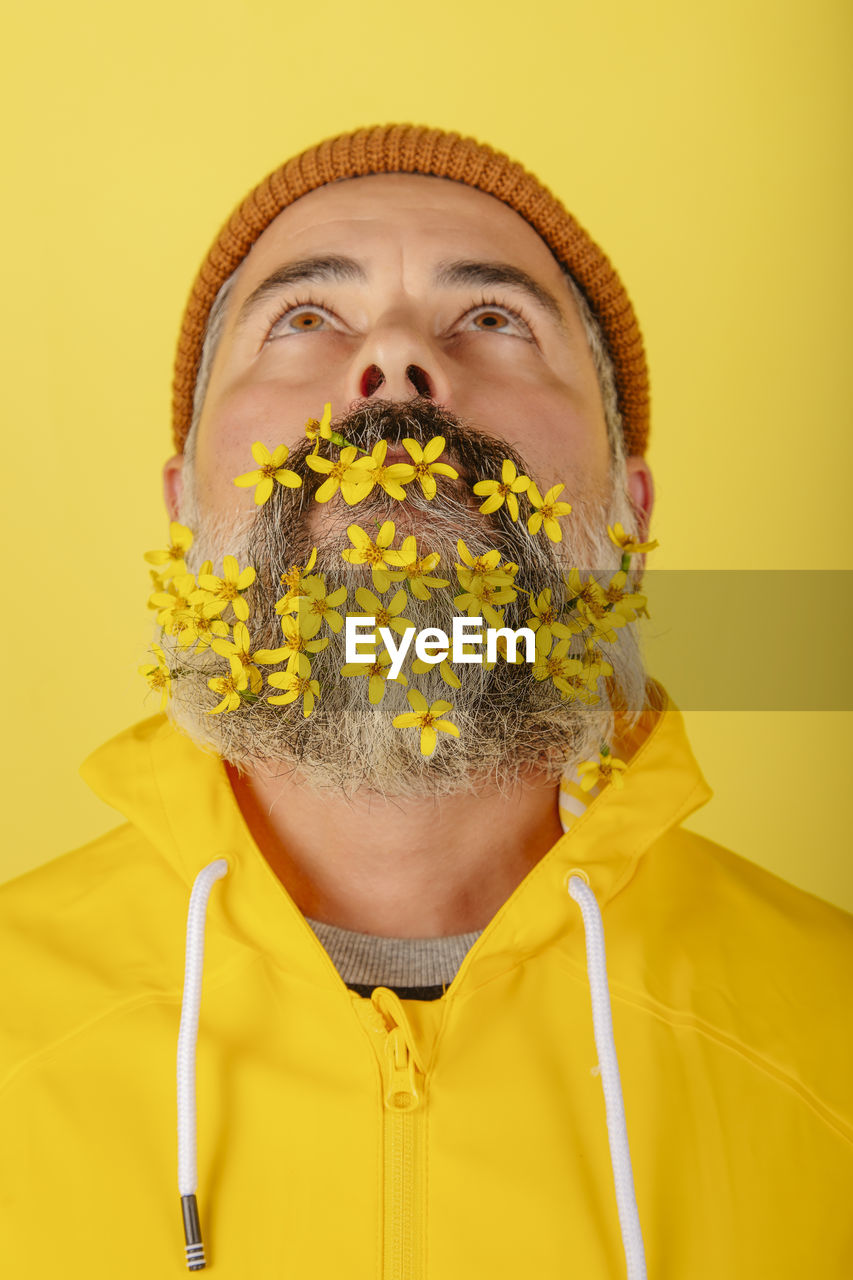 Close-up of man with flowers in his beard, standing against a yellow background looks up. 