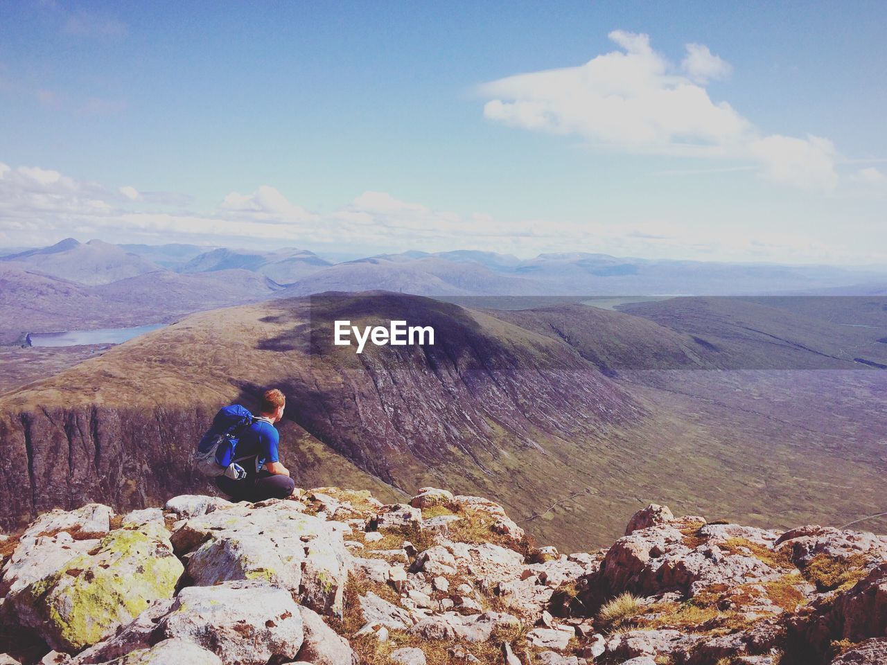 Rear view of male hiker crouching on rock at mountain against sky