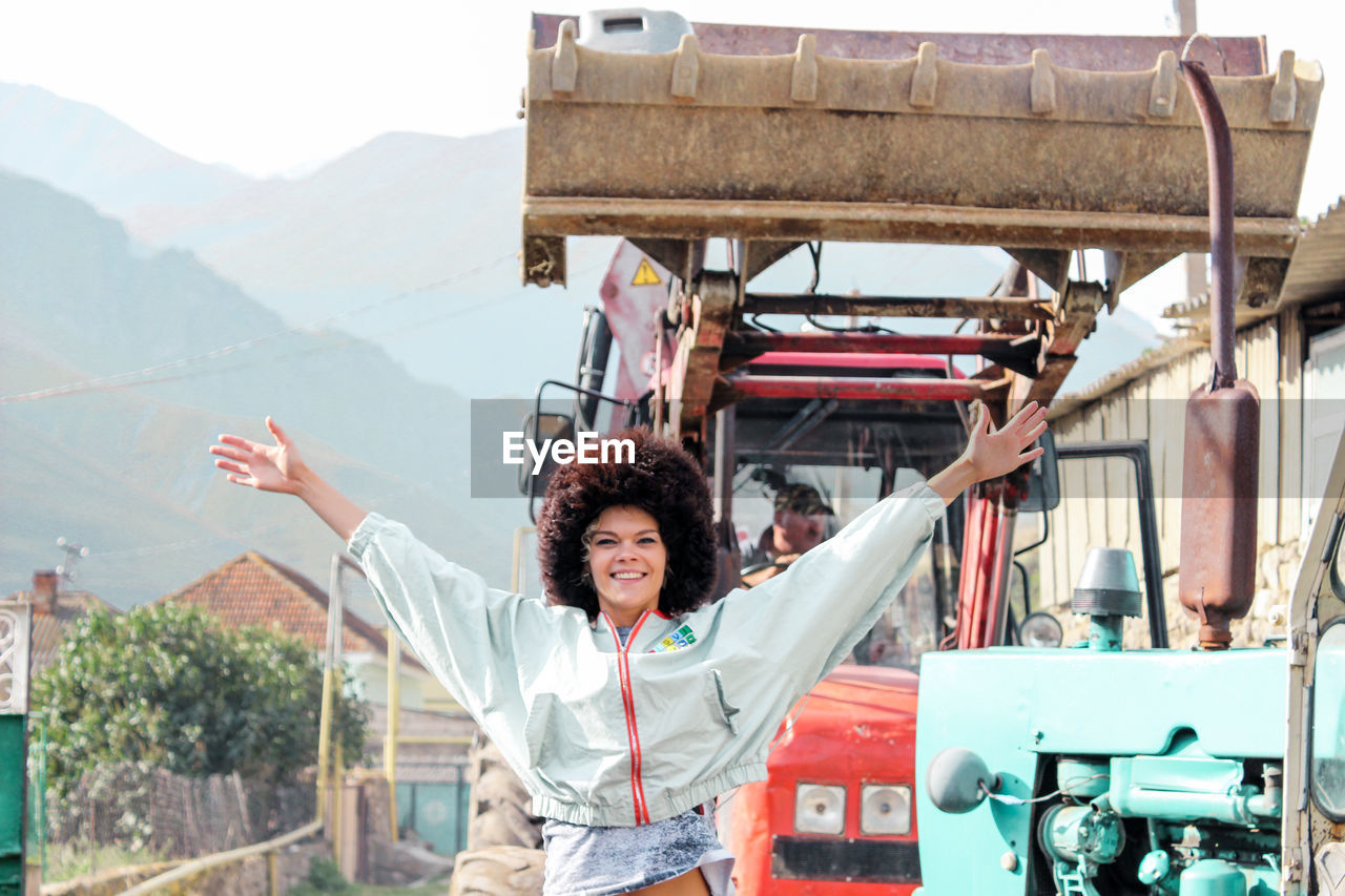 Portrait of happy young woman with arms outstretched standing against bulldozer