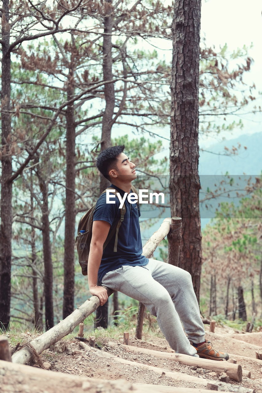Side view of young man sitting on railing in forest