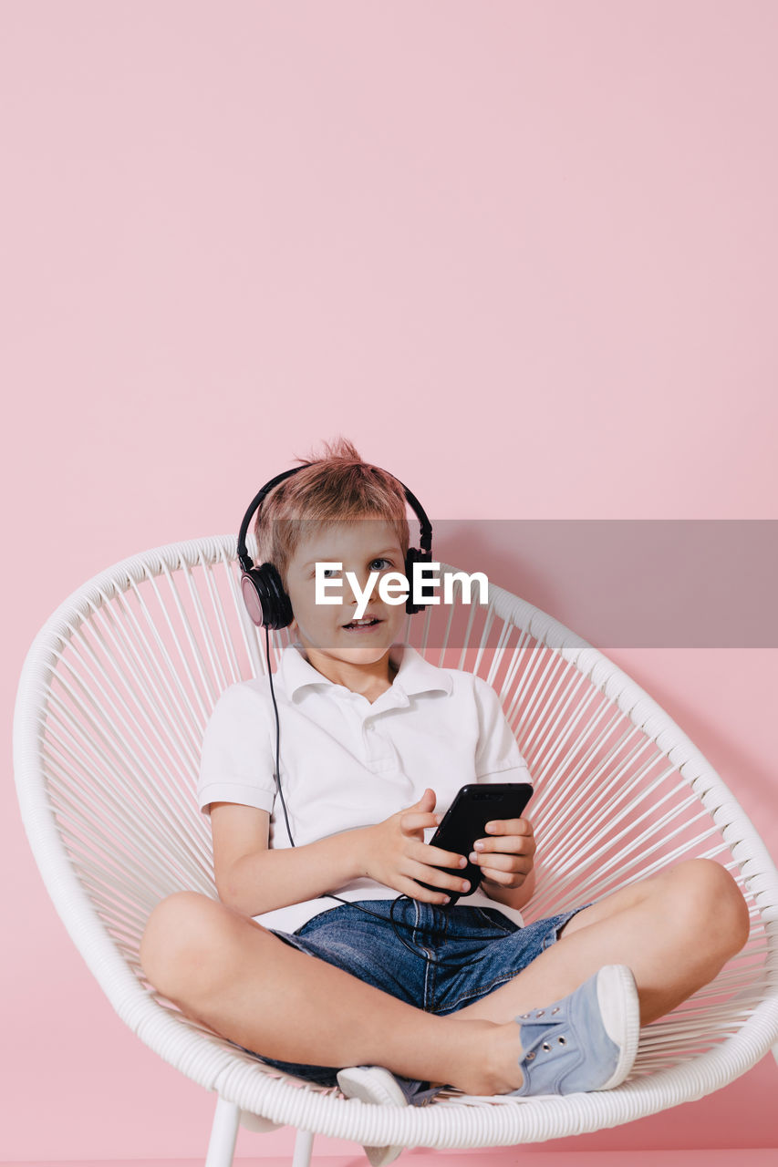 Portrait of boy listening music while using phone on chair against colored background