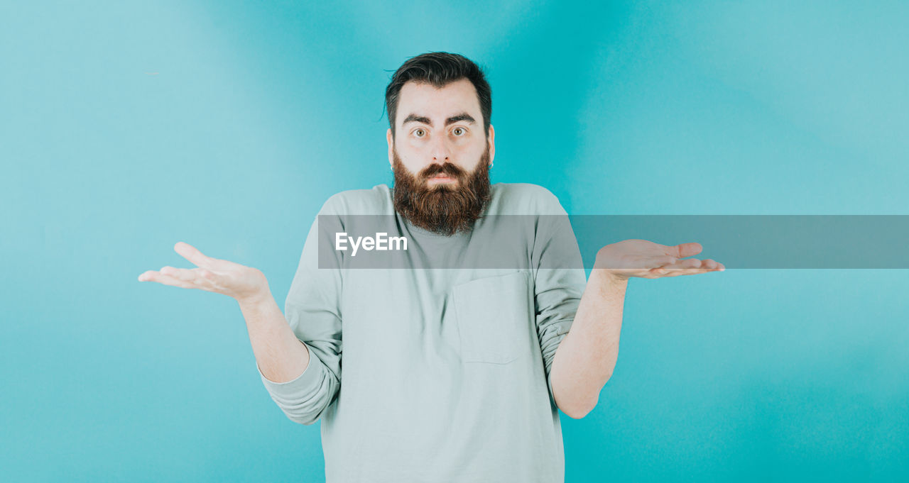 one person, adult, beard, facial hair, colored background, blue, studio shot, men, gesturing, indoors, waist up, blue background, portrait, person, front view, sign language, standing, copy space, young adult, emotion, clothing, hand, looking at camera, casual clothing, human face, finger, arm, looking, limb, lifestyles, fun