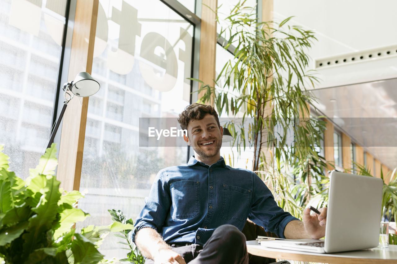 Cheerful young businessman looking away while sitting with laptop at table in cafe