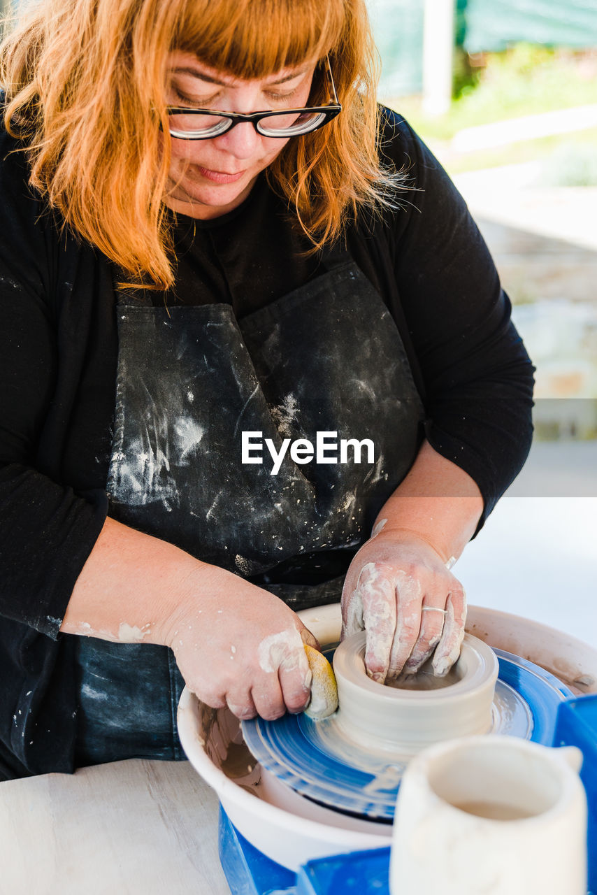 Focused woman standing near table with throwing wheel while sculpturing with clay in workshop