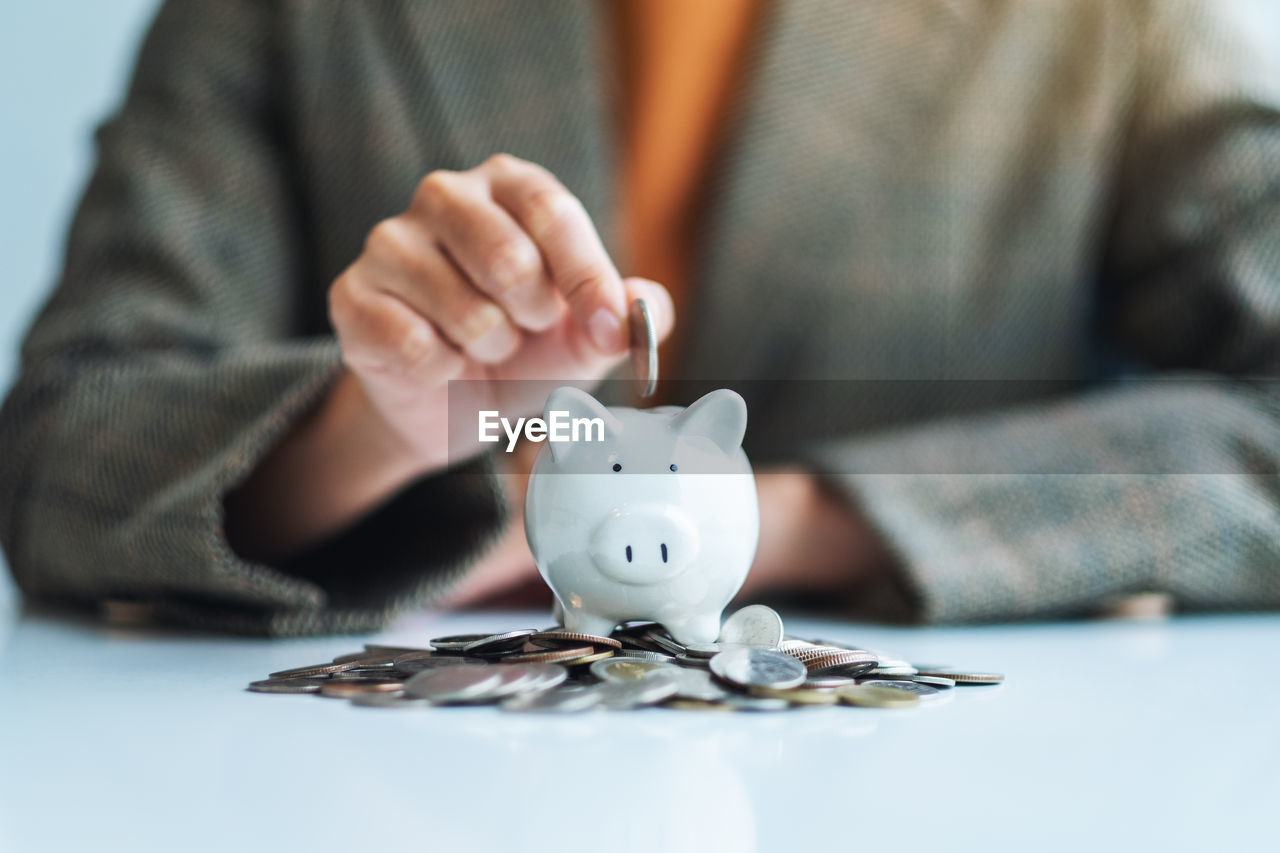 midsection of woman inserting coin in piggy bank on table