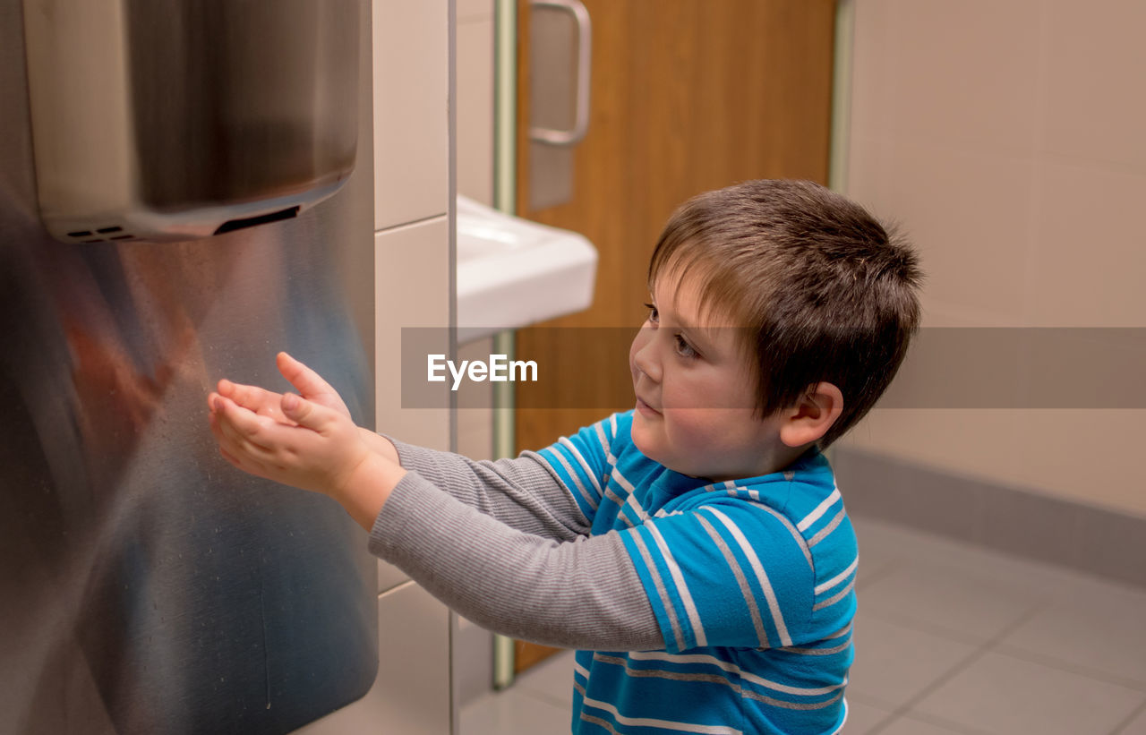 A young boy uses a air hand dryer to dry his hands after washing them in the bathroom sink.