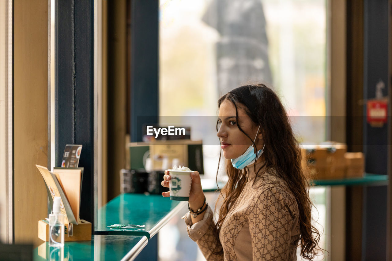 A beautiful teenage girl drinking coffee in bar wearing medical mask