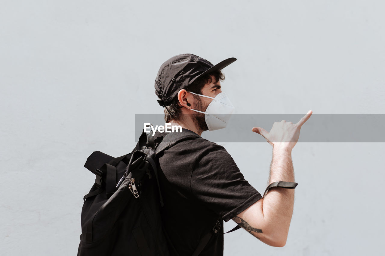 Back view of unrecognizable trendy man with backpack in respiratory mask demonstrating shaka gesture near wall while looking away during coronavirus pandemic