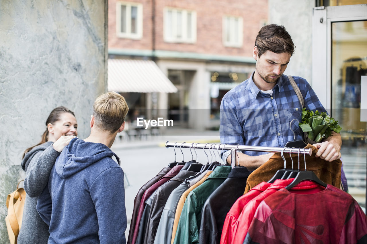 Young man selecting jackets with friends passing by clothing store