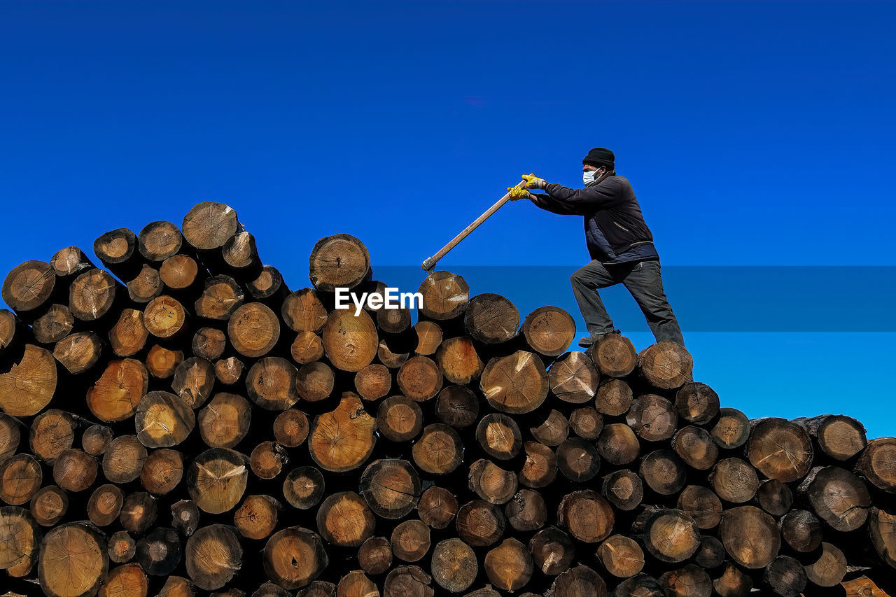 Low angle view of man standing on logs against clear blue sky