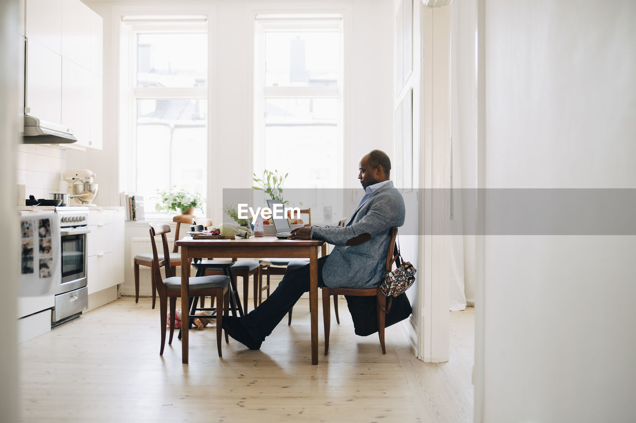 Full length of mature man working on laptop while sitting in kitchen at home