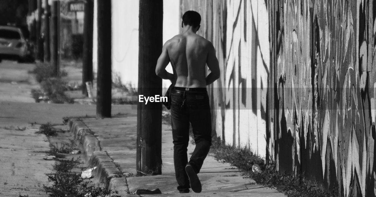 Rear view of shirtless man walking on footpath by wall with graffiti