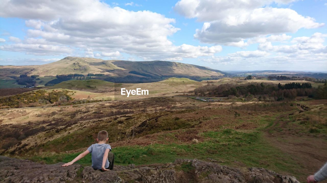 Full length of boy sitting on rock formation against cloudy sky