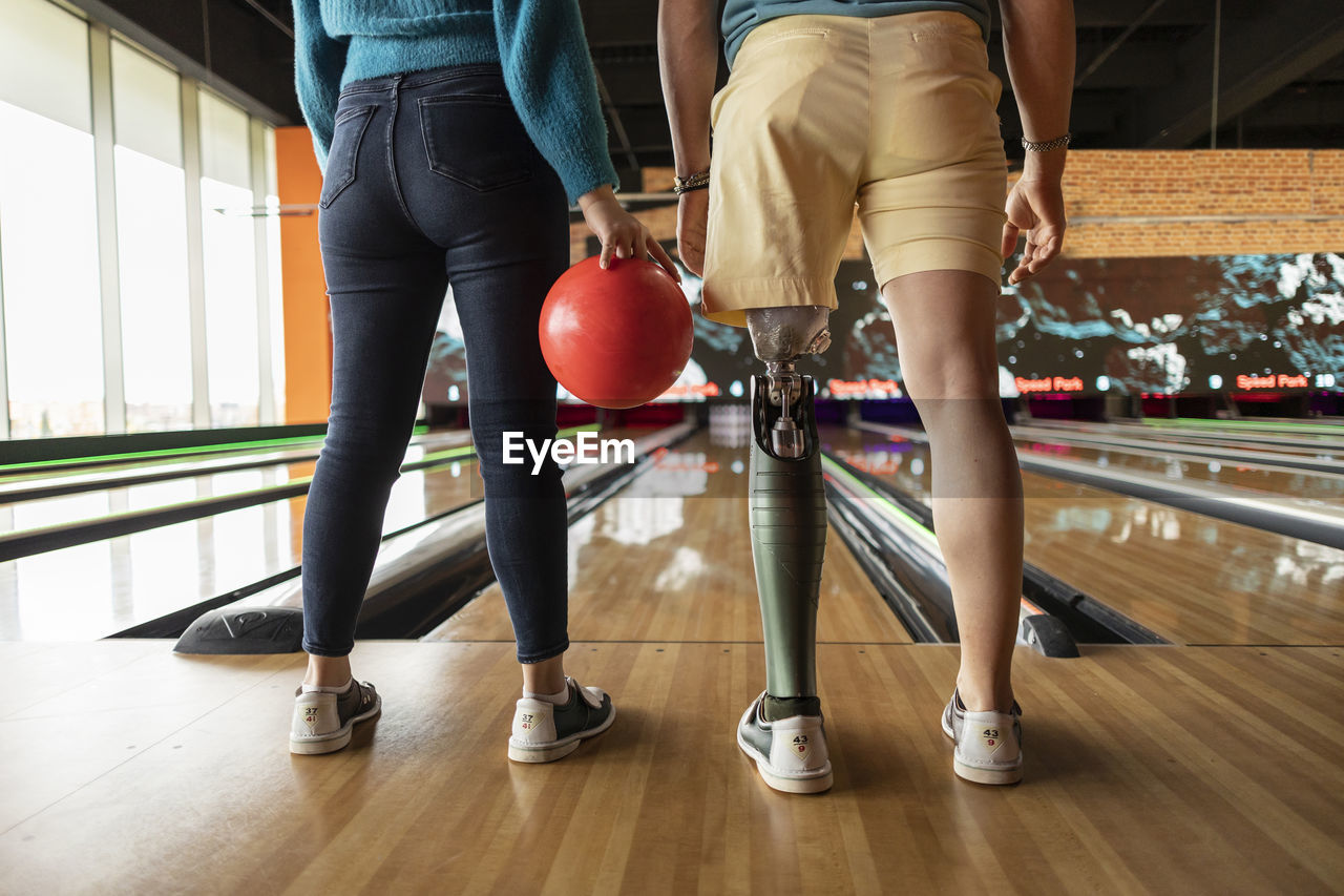 Woman holding ball with friend wearing artificial limb standing on hardwood floor at bowling alley