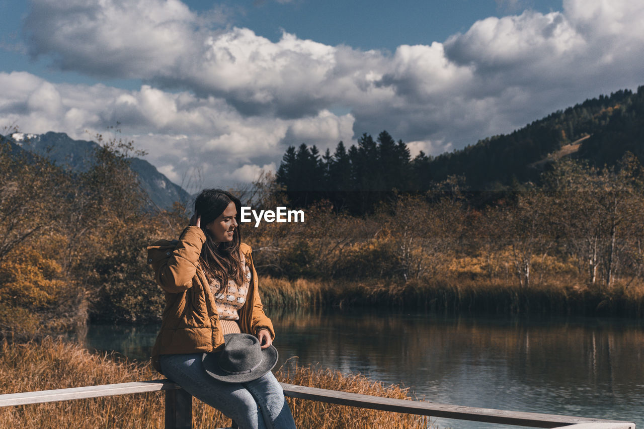 Young woman sitting on fence on shore of lake in autumn