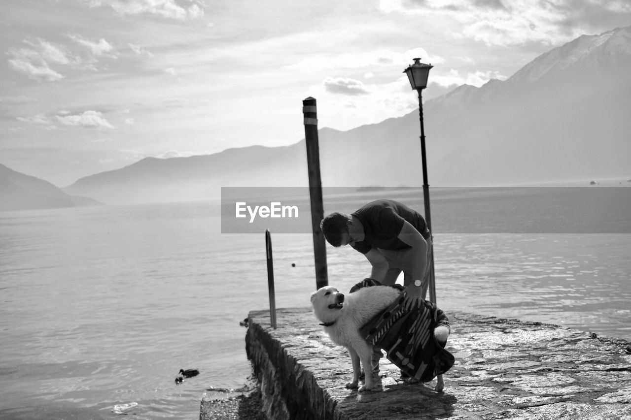 Man wiping dog with towel on pier over lake against mountains