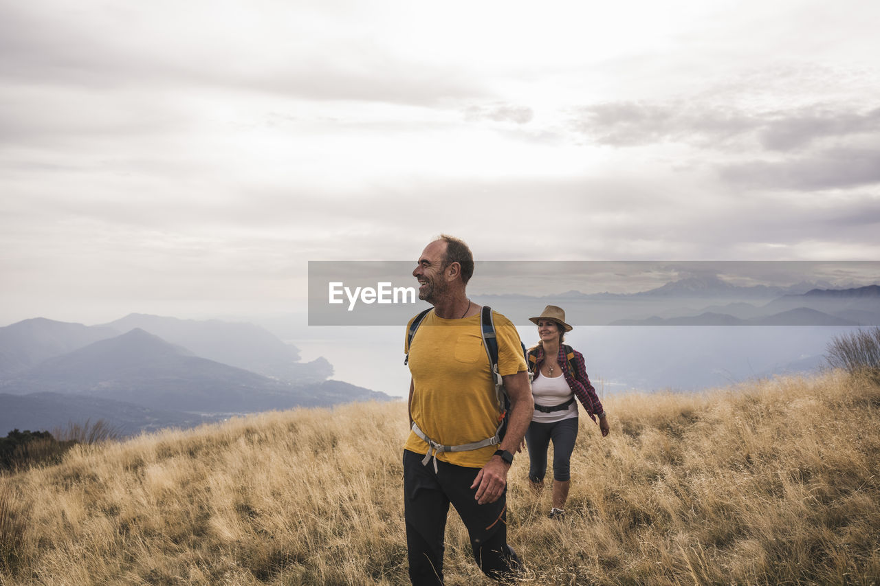Happy mature man and woman hiking on mountain under cloudy sky